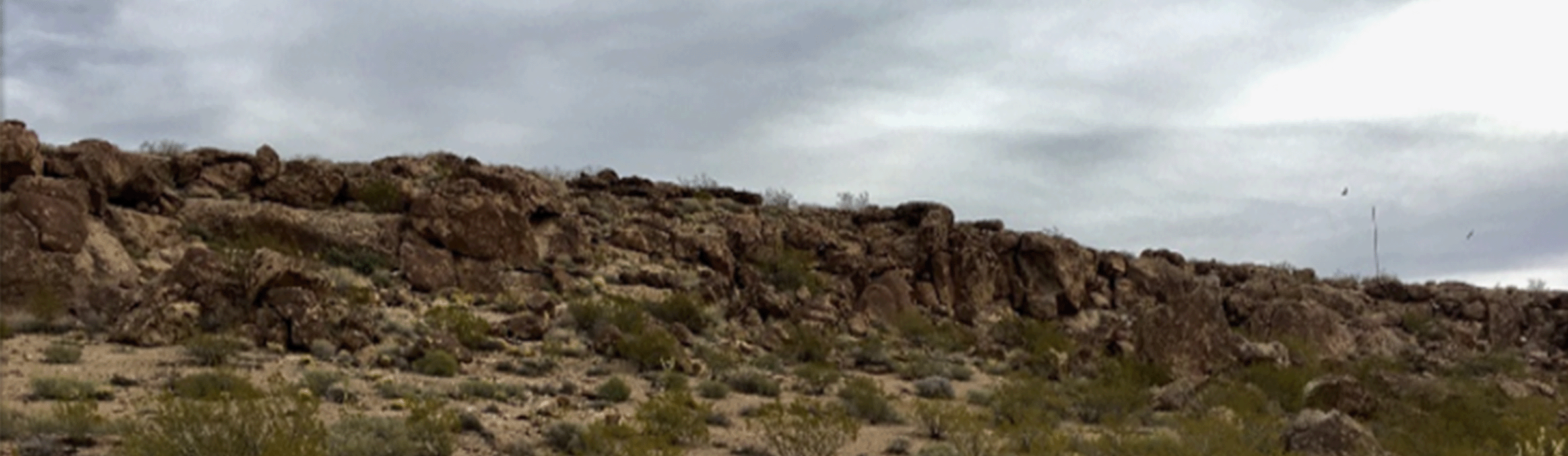 Panoramic photo of a rocky ridge stretching across a desert landscape with sparse green shrubs under a cloudy sky at the Arizona Army National Guard Kingman Range MRS